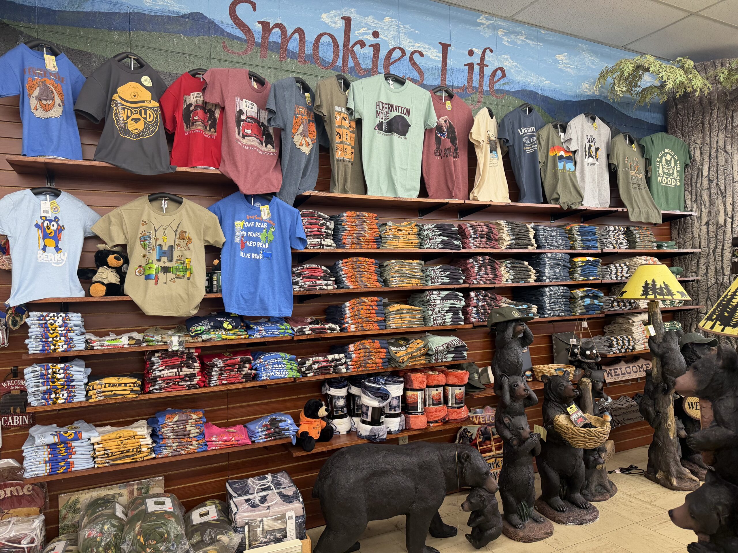Wall display of Smokies Life T-shirts, Smoky Mountain-themed apparel, blankets, and black bear décor inside a gift shop. Shelves feature colorful graphic tees inspired by the Great Smoky Mountains and Tennessee wildlife.